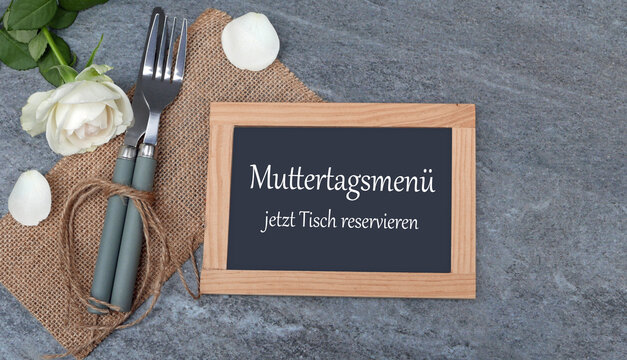Kleine Tafel mit der Aufschrift 'Muttertagsmen&uuml; jetzt Tisch reservieren' neben Besteck auf grauem Untergrund mit wei&szlig;en Rosen.