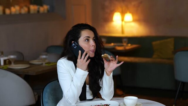 Happy pretty lady talking on mobile phone while biting bruschetta during lunch in modern restaurant. Copy space. Business, lifestyle concept. Real time video.
