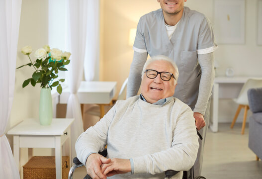 Smiling elderly man in wheelchair enjoying time at home with kind caregiver standing behind him. Happy senior expressing positivity and gratitude, receiving care and support during rehabilitation.