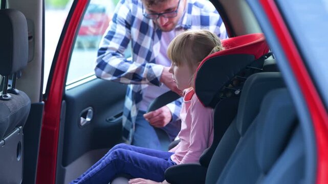 Young father helping her daughter sitting in the car seat while fastening her with a seat belt. Family, transport, trip, safety and parenthood concept. Real time