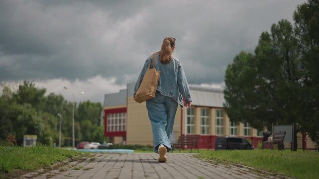 Woman walking toward camera storm clouds, low angle urban building backdrop, confident stride with denim jacket and tote bag, dramatic sky and moody light, purposeful commute, cinematic perspective