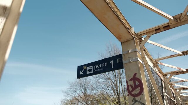 A blue informational sign for platform one hanging from an old metal bridge structure against a clear sky