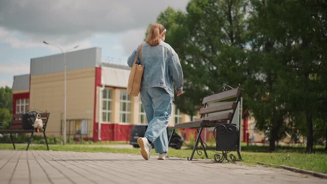 White woman walking in urban park framed by modern school building and tall trees casual denim outfit and tote bag pauses to sit on bench checks phone breathes in sunlight conveying relaxed thoughtful