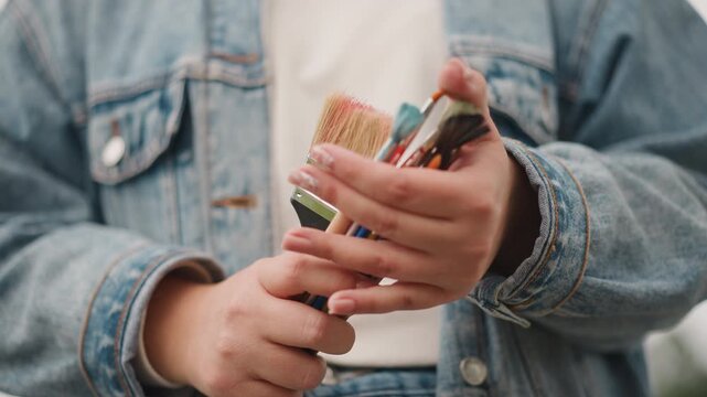 Closeup hands organizing mixed brush sizes in denim jacket, artist prepping tools for canvas or mural, steady grip and ferrule focus, routine workflow, outdoor daylight, practical preparation