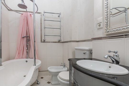 A small bathroom includes a bathtub with a shower and a pink curtain. The sink and toilet are also in the room, showing clean lines and simple design. Light comes in from outside