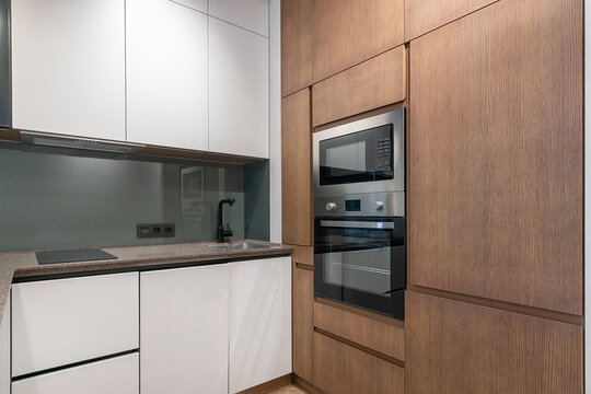 A kitchen features wooden cabinets and stainless steel appliances. The space includes a sink, cooktop, and double oven. Bright light fills the room, showing a clean and organized look