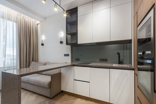 A kitchen and living area are combined in a compact space. Natural light enters through large windows, illuminating the countertops and sofa