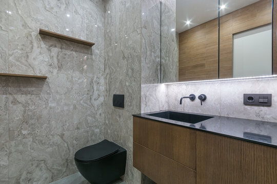 A bathroom is shown with marble tiles on the walls, a black toilet, and a dark sink. Wooden shelves are mounted on one wall. Bright lights are installed in the ceiling