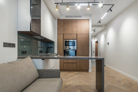The scene shows a kitchen and living area in an apartment. The kitchen features wooden cabinets and modern appliances. A couch is placed nearby, creating a cozy space for relaxing or dining