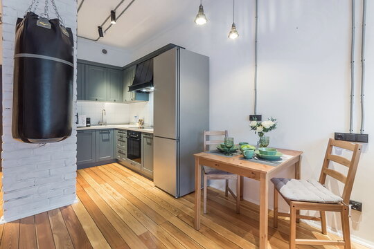 In a small apartment, a kitchen area is seen with a punching bag hanging nearby. A wooden dining table is set for two with dishes and a plant. Light comes in from the windows