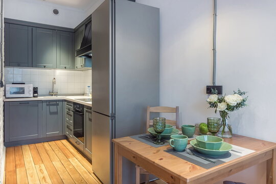 A small kitchen and dining space shows a table with green dishes and glasses. The kitchen has a fridge and a microwave. Sunlight enters the room through a window