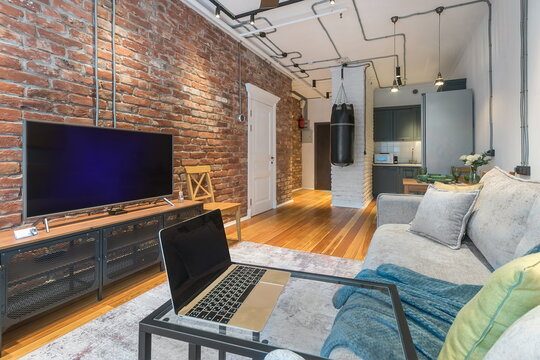 A cozy apartment features a living room with a brick wall and a television. A laptop rests on a table. The kitchen can be seen in the background, showing modern design