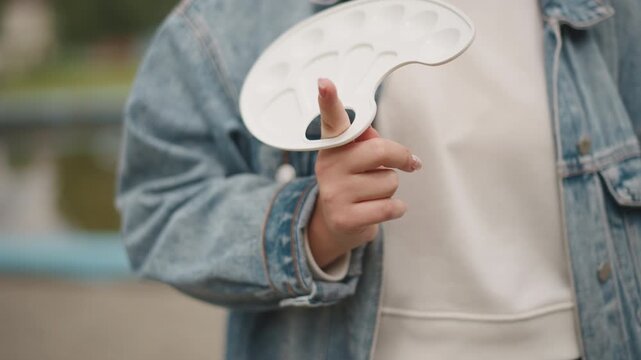 Closeup artist holding paint palette outdoors denim jacket white hoodie finger through palette hole empty white mixing wells soft daylight park railing background calm preparation for painting casual