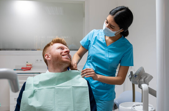 Dentist in mask and scrubs preparing happy male patient for a dental checkup in clinic