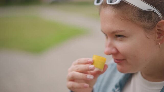 Young caucasian woman sniffing yellow cup, park closeup, sunglasses on head, gentle smile, thoughtful expression, handheld sample container, natural light, soft background, leisure moment