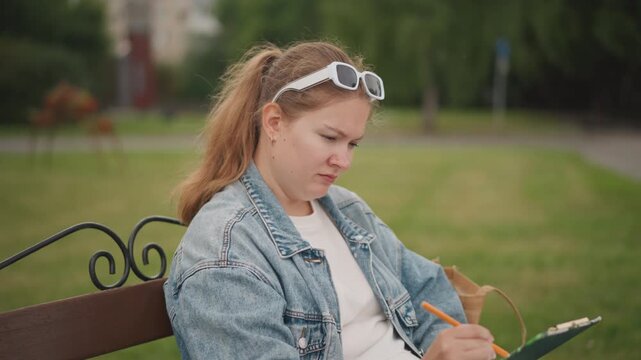 Young woman focused on clipboard sketching composition, denim jacket and sunglasses headband, portfolio bag and notes at side, concentrated expression, urban green space with walkway and trees,