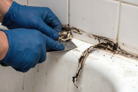Worker removing moldy silicone sealant from bath, close-up of hands with blade, renovation prep detail