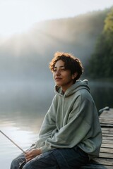 Fototapeta premium Young person sitting relaxed holding fishing rod on dock at misty lake