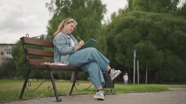 Caucasian artist taking plein air notes on bench sketchbook and pencil in hand, denim outfit and sneakers, urban trees and pathway backdrop, concentrated expression capturing quick studies