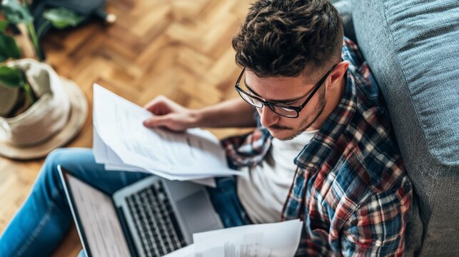 Young man remote working at home, focused on reading documents and reviewing paperwork while using a laptop for productivity in a cozy environment
