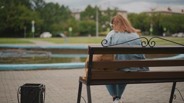 White woman sitting on bench by fountain, denim jacket, rolling suitcase at feet, urban park background with trees and distant buildings, overcast sky, reflective mood, slow cinematic pan