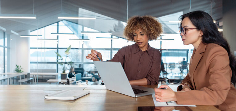 Two professional women, businesswomen in modern open-plan office workspace collaborating at laptop, discussing business work on computer pc screen and taking notes at a desk. Web banner, copy space