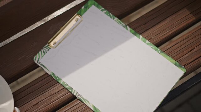 Empty clipboard on wooden bench greentrim metal clip casts diagonal shadow across blank recycled paper beside ceramic cup on slatted patio table warm wood grain and soft morning sunlight creating calm