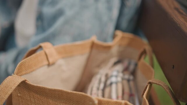 Closeup jute tote on bench with checked cloth, street artist packing supplies, denim sleeve reaching into bag, candid urban scene, textured fibers, daylight bokeh, casual commute vibe