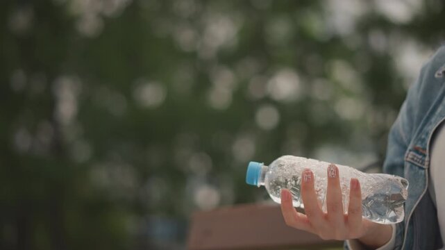 Woman on park bench fidgets with water bottle, denim jacket, playful wrist motions and cap spinning, candid moment of nervous energy turned mindful, soft treelined background and relaxed vibe.