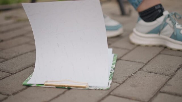 Blank notebook flipping on pavement, teal sneakers and denim ankles visible, page curls from breeze, clipboard base resting on textured brick, soft sunlight and shadow, candid street detail