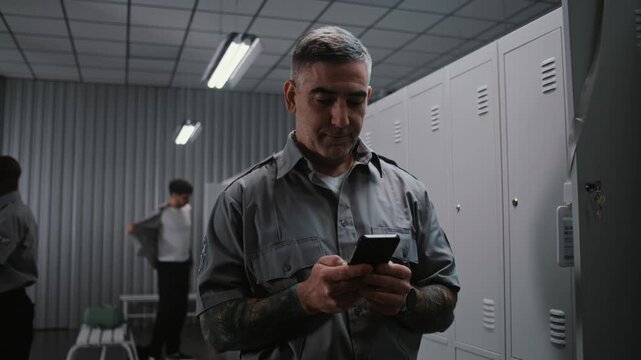 Security Officer With Arm Tattoos Checks Notifications and Social Media on Smartphone by Locker, While Other Staff Members Undress After Airport Shift. Workplace Communication and Relaxation Concept.