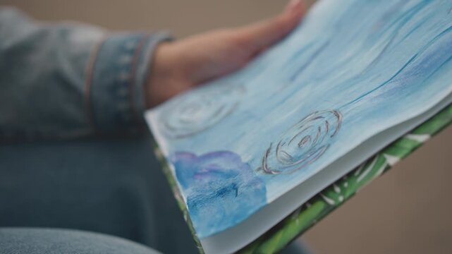 Hands painting blue seascape on canvas, denim sleeve visible, delicate brushwork creating wave textures and spiral shell detail, soft natural light, closeup of meditative creative process