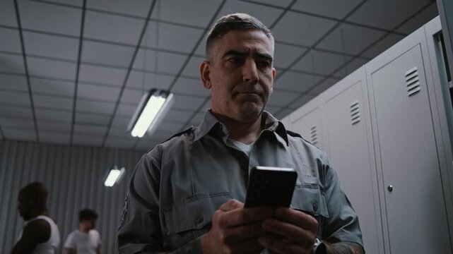 Serious Middle Aged Male Security Officer With Prominent Arm Tattoos Stands by Grey Metal Locker, Concentratedly Typing Message or Email Response on Smartphone After Finishing Airport Security Shift.
