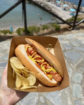  Hot dog with ketchup and mustard served with potato chips in a takeaway box, held by hand with a seaside view in the background. Street food by the beach, sunny summer day, casual outdoor eating.