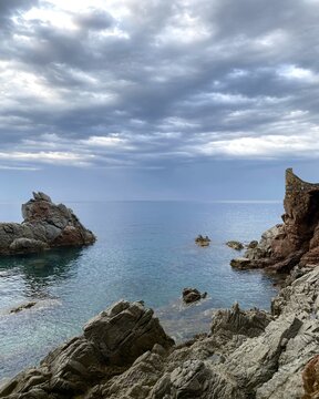  Rocky coastline with calm sea under dramatic cloudy sky. Clear blue water surrounded by rugged cliffs, creating a peaceful yet moody atmosphere. Natural seascape with soft light and horizon view.