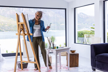Middle-aged female artist standing and painting in home studio with easel, palette, headphones © wavebreak3