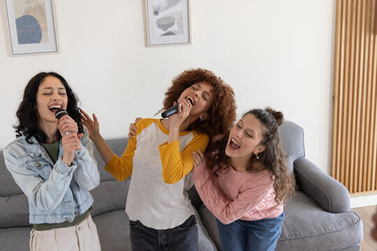 Diverse female friends singing into wireless microphones, leaning in living room near gray sofa