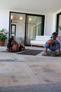 African American family doing yoga on patio on yoga mats parents holding plank child climbing back