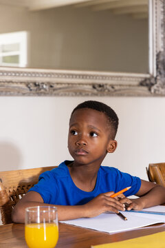 African American school-age boy sitting at dining table in blue tee holding orange pencil, notebook