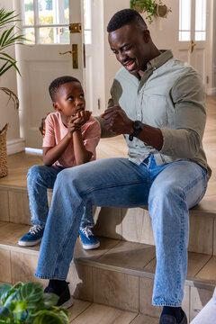 African American father and son sitting on wooden steps at home, talking near glass door