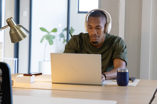 African American man typing on silver laptop at wood desk wearing headphones with tumbler nearby