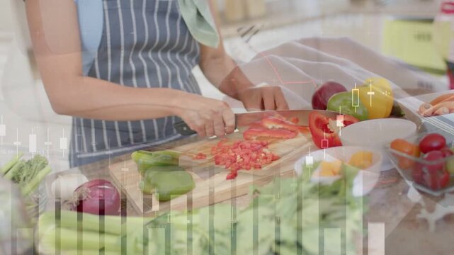 Woman starting slicing red pepper on board for meal prep while finance charts overlaying hands