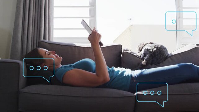 Woman holding tablet on sofa checking messages, new chat bubbles floating near head and dog tech