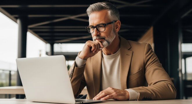 Professional man with grey hair works thoughtfully on his laptop in a modern outdoor setting.