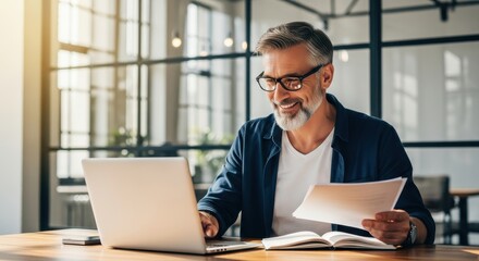 Obraz premium Mature man working on a laptop at an office desk while holding a document.