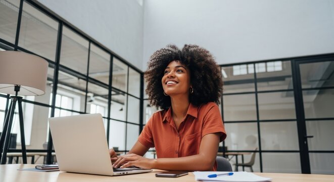 Young professional woman with curly hair smiles while working on her laptop in a modern office.