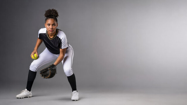 Focused softball player in a low defensive stance grips a yellow ball and open glove against a gray studio backdrop with wide copy space.