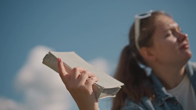 Outdoor woman joyfully displays her book. Lady dressed in denim celebrates freedom through reading outdoors. Woman in casual denim exudes happiness as she proudly elevates her book outside