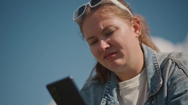 Young woman outdoors reading phone frustrated, denim jacket and sunglasses on head under bright blue sky closeup portrait shows squinting and frown while checking messages, candid cinematic lighting,