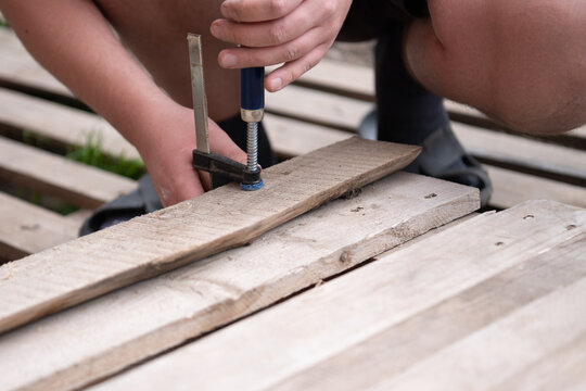 A close-up shot showing a person's hands tightening a metal F-clamp on a wooden board. The process of manual woodworking, DIY project, or furniture repair. Focused view on the tool 